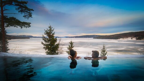 Une piscine à débordement au fond de verre offrant une vue sur un lac gelé et des arbres.