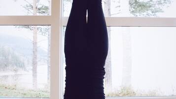 Una persona haciendo un pino sobre una colchoneta de yoga morada en una habitación con ventanas amplias, suelo de madera y velas en el suelo.