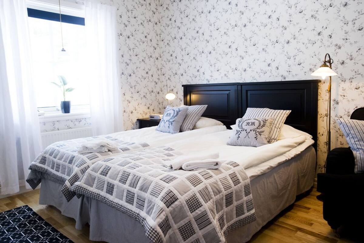 A neatly made bed with a checkered bedspread, a bedside lamp, and a window with a potted plant.