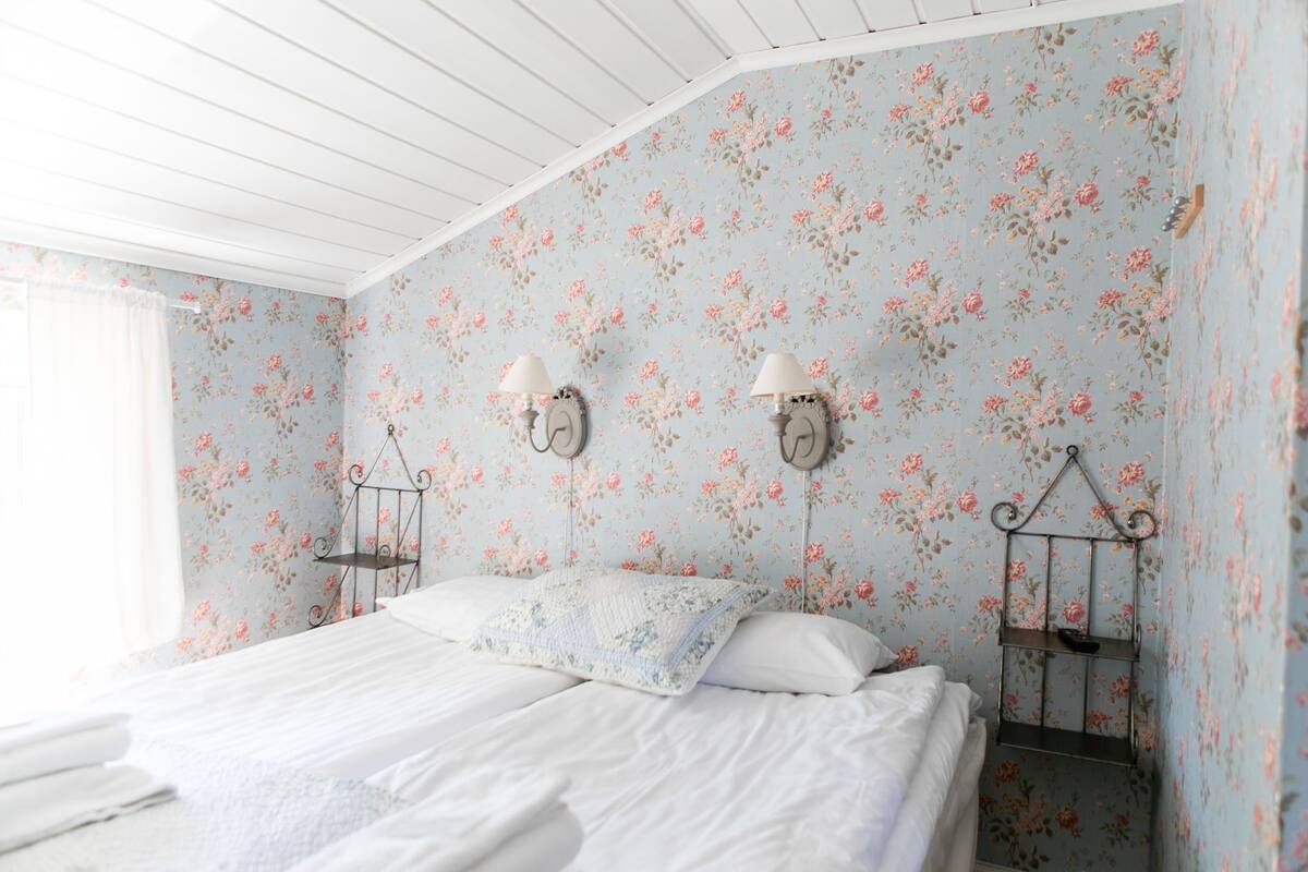 A bedroom with floral wallpaper, two bedside lamps, a bed with white linens, and a hanging shelf.