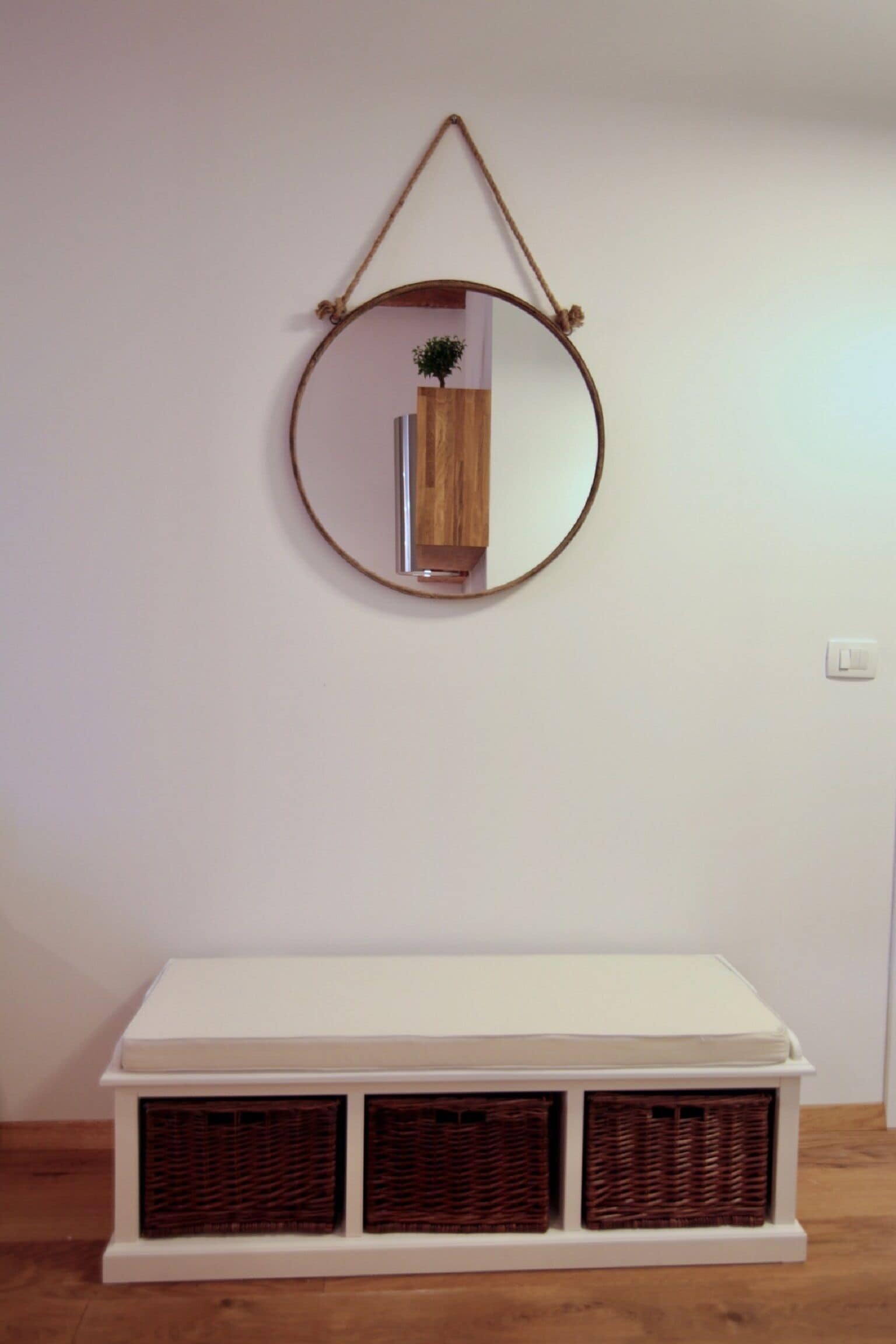 A white console table with three wicker baskets underneath, a round mirror hanging on the wall above, and a small wooden plant in the mirror's reflection.