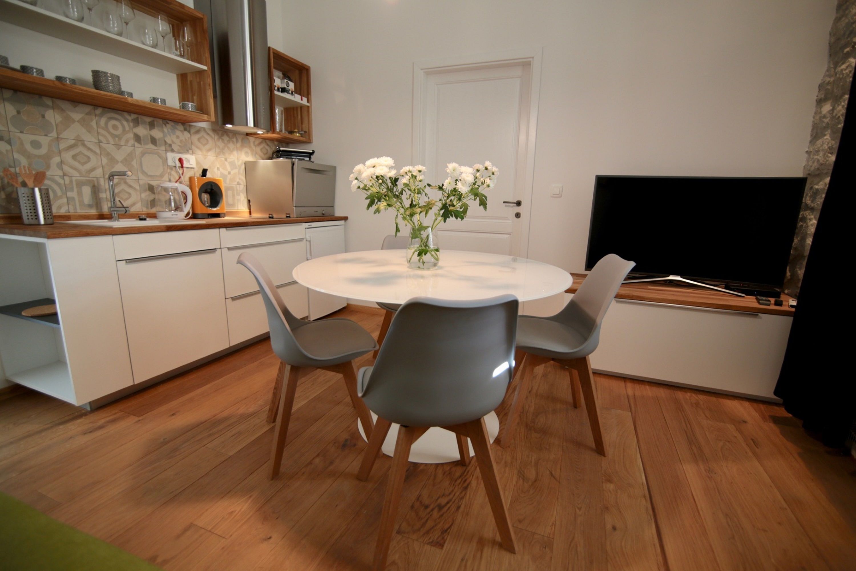 Une cuisine moderne avec des armoires blanches, un plan de travail en bois et un four encastré. On y trouve une table à manger avec quatre chaises et un vase rempli de fleurs blanches. Un téléviseur à écran plat est fixé au mur.