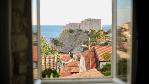 Un pueblo costero con un imponente castillo de piedra que domina el mar, rodeado de edificios escalonados con tejados de tejas rojas.