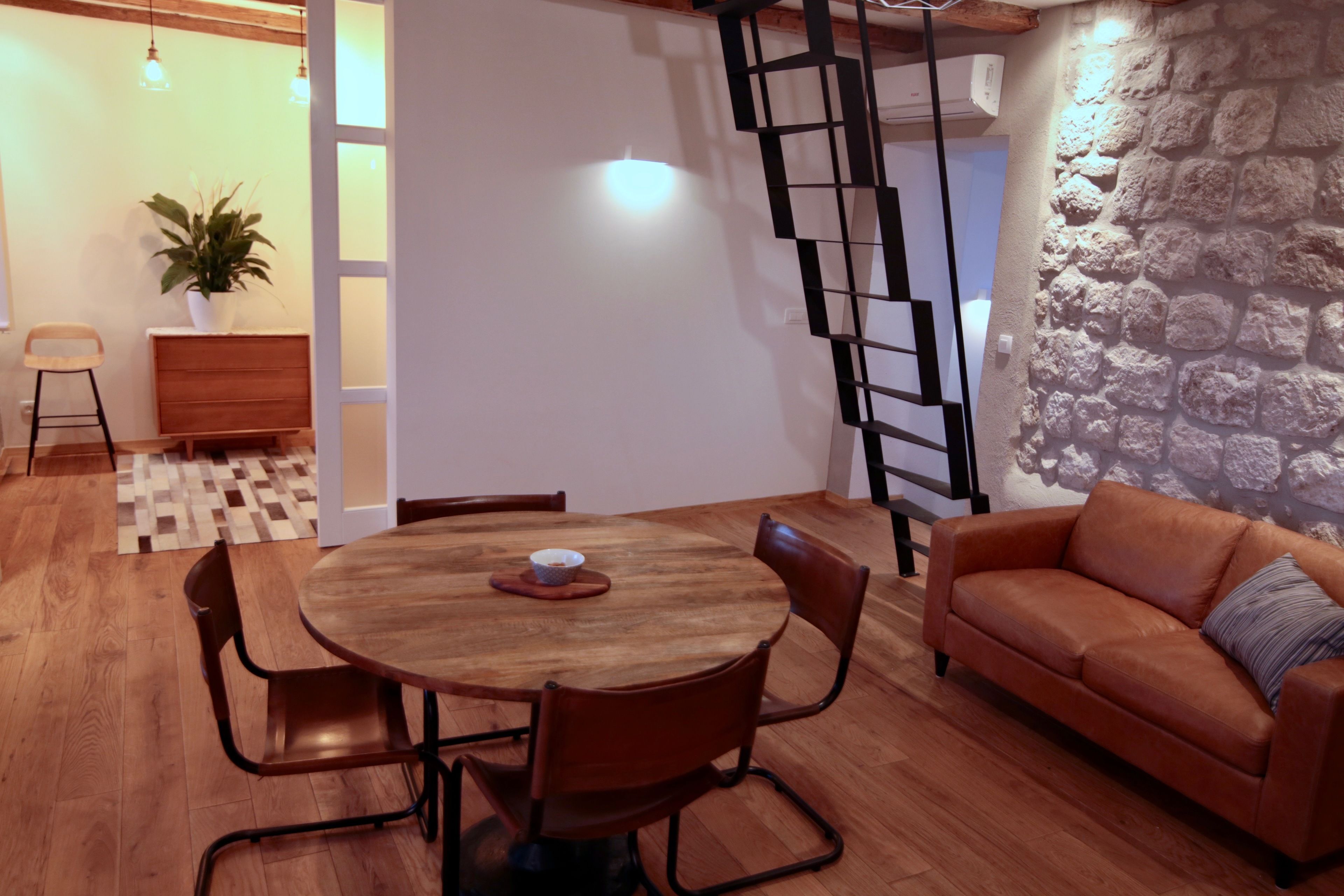 Ein Essbereich mit Holztisch und -stühlen, einer Couch, einer Zimmerpflanze und einer Treppe mit schwarzer Geländer.