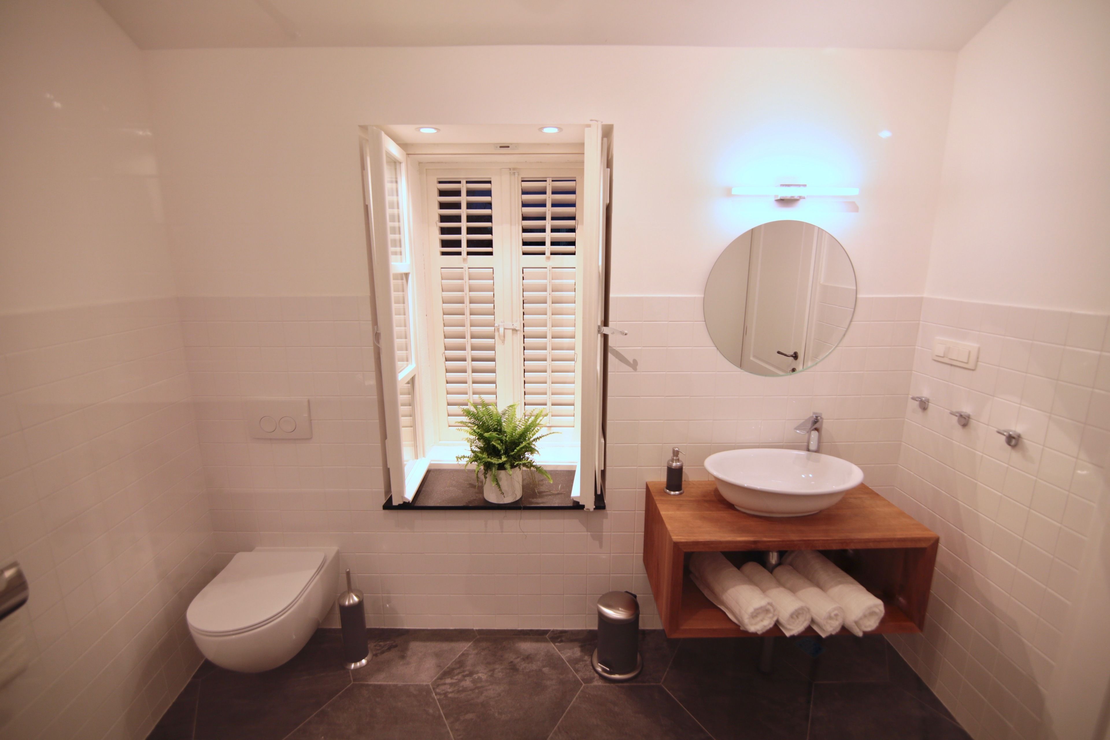 A bathroom with a toilet, sink, mirror, and a window with shutters.