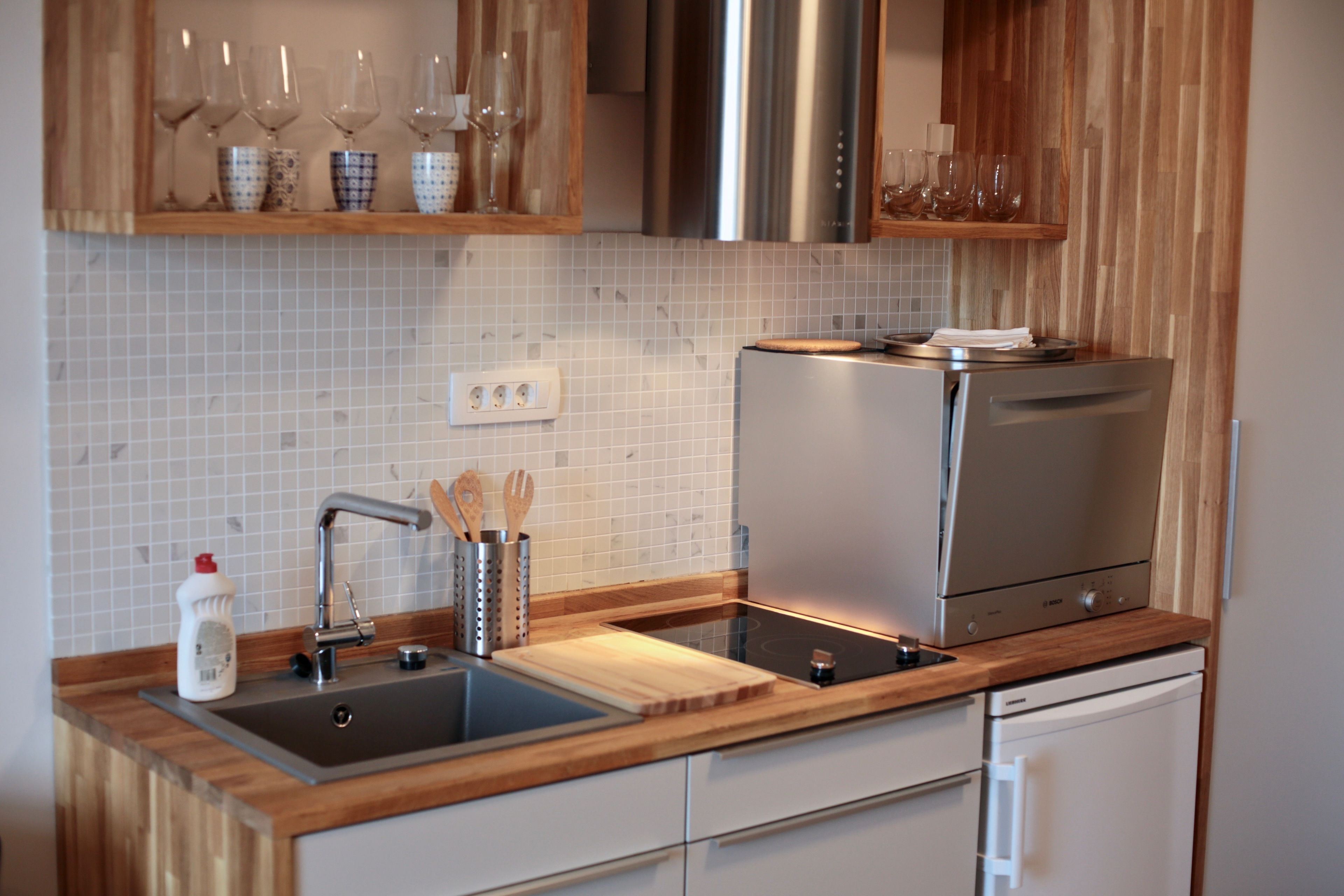 A modern kitchen with a stainless steel refrigerator, a built-in oven, a sink with a wooden countertop, and a backsplash of white tiles.