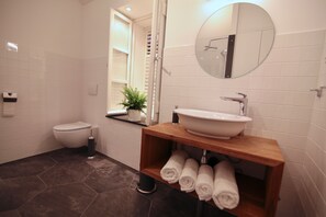 Ein modernes Badezimmer mit Toilette, einem Holzwaschtisch mit Waschbecken und einem Spiegel darüber. Handtücher liegen auf dem Regal und ein Topf mit einer Pflanze steht auf der Ablagefläche.