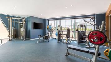Un gimnasio con diversos equipos de ejercicio, amplias ventanas y una pared azul.