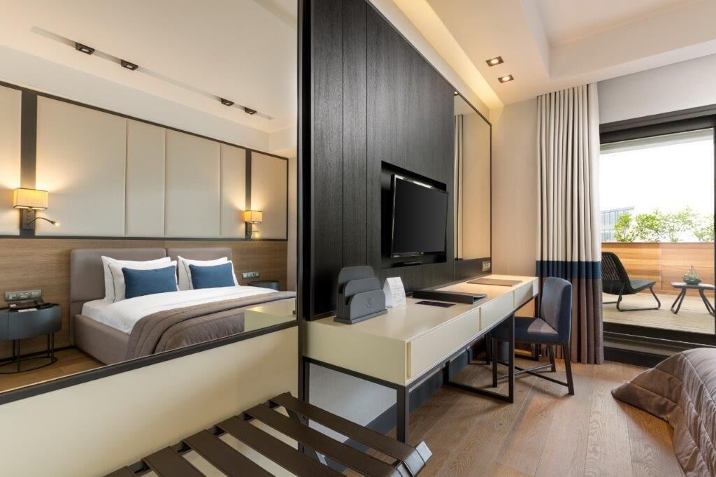 A modern hotel room with a bed, desk, chair, TV, and a view of the outdoors.