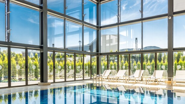 Une grande piscine intérieure aux parois en verre transparent, entourée de chaises longues et offrant une vue sur la verdure environnante.