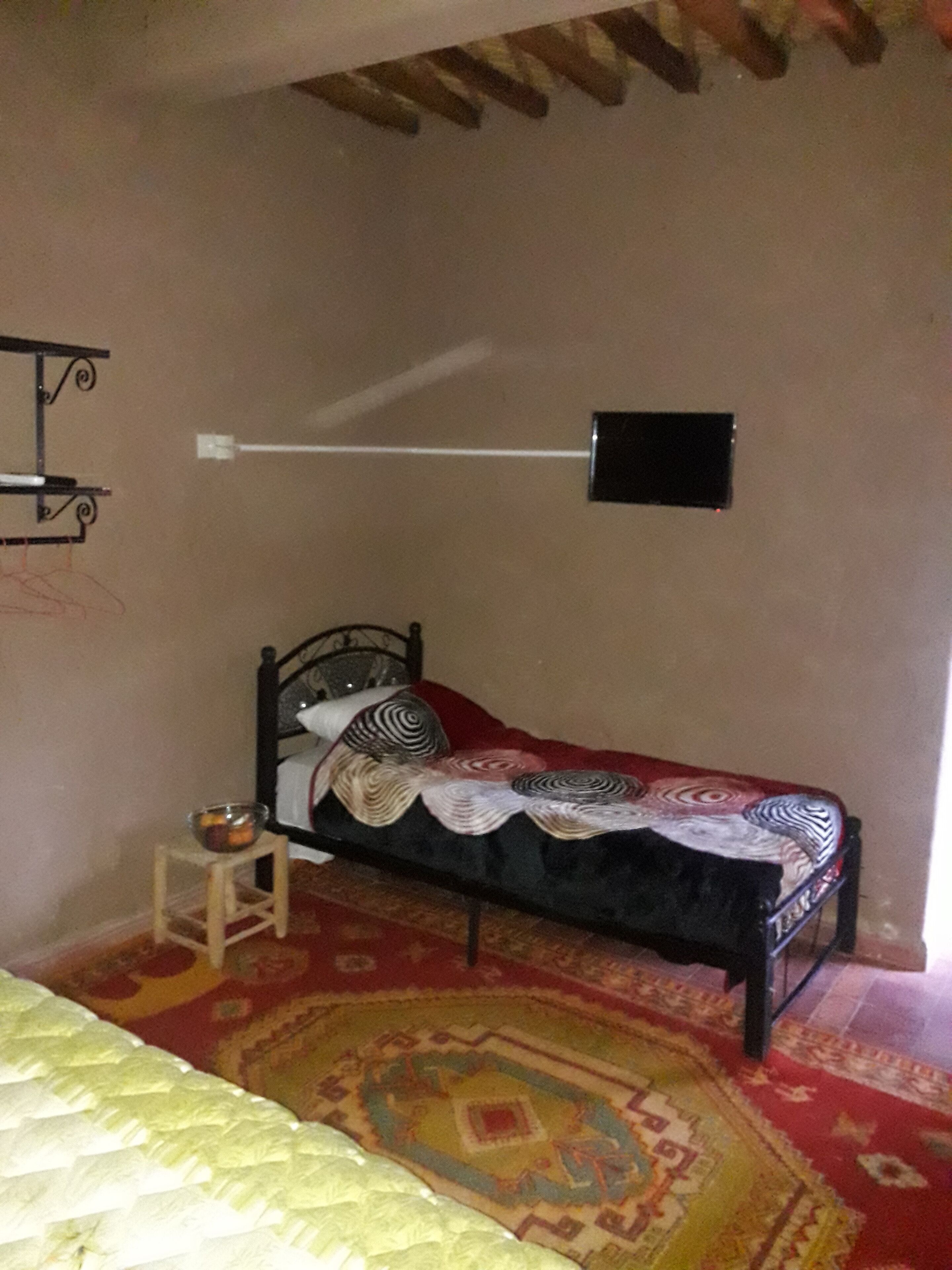 Ein Schlafzimmer mit einem Einzelbett, einem kleinen Tisch mit einer Obstschale, einem gemusterten Teppich und einem an der Wand befestigten Fernseher.