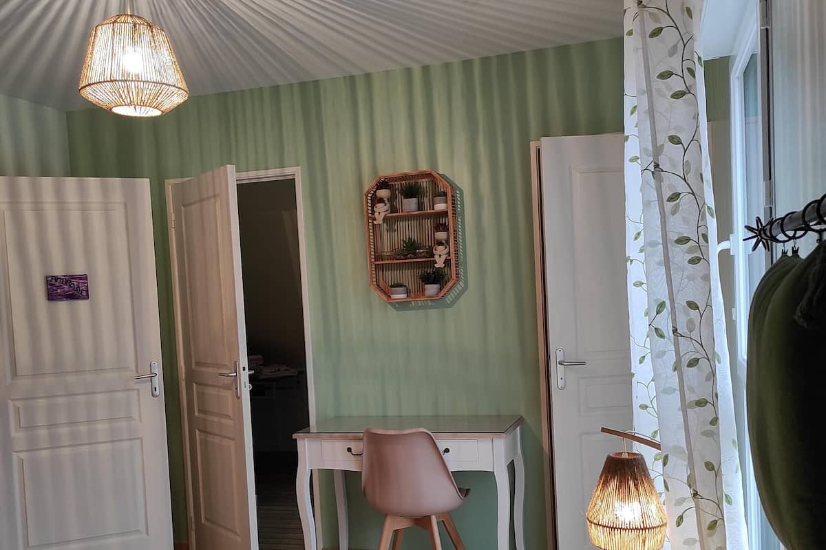 A room with a bed, a desk, and a chair. There is a ceiling fan, a wall-mounted shelf with plants, and a window with curtains.