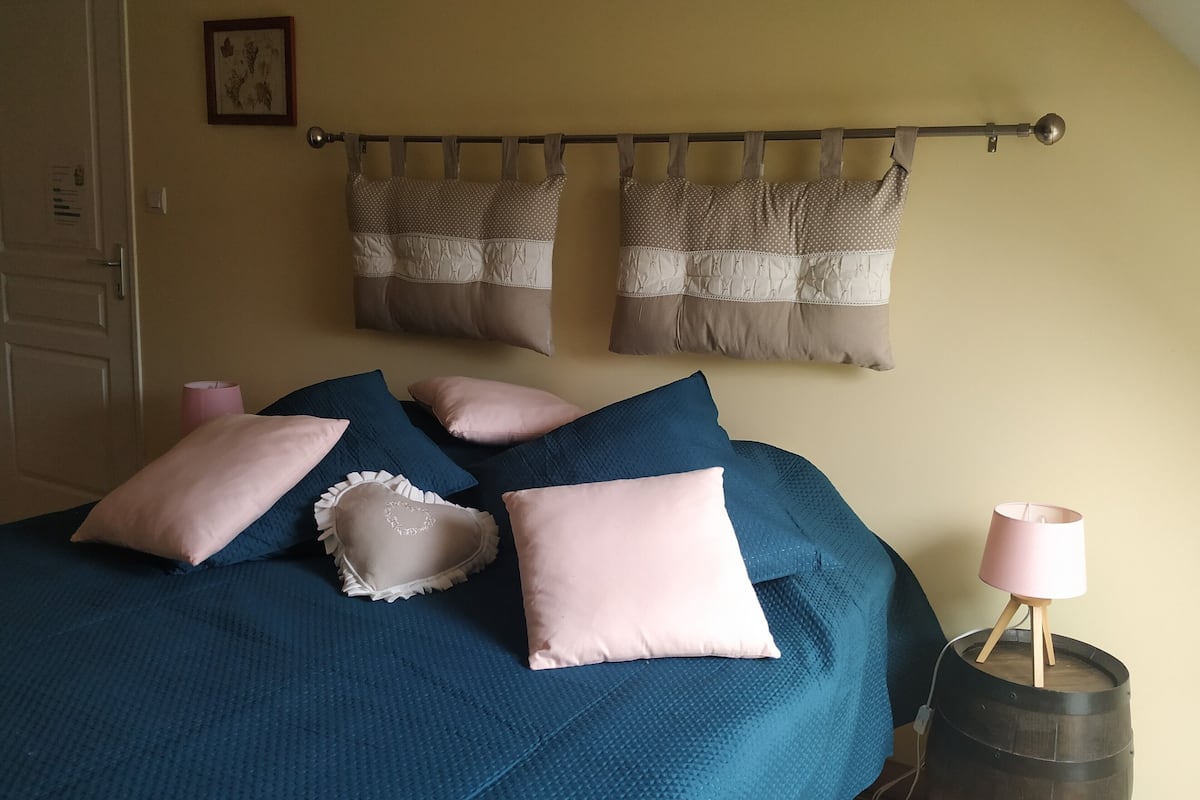 A bedroom with a bed covered in teal bedding, multiple pillows, a wooden barrel, and a curtain rod with two curtains.