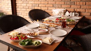 A dining table set with various dishes, plates, and cutlery, surrounded by wicker chairs, against a brick wall. - Mandalin Pansiyon (Seferihisar)