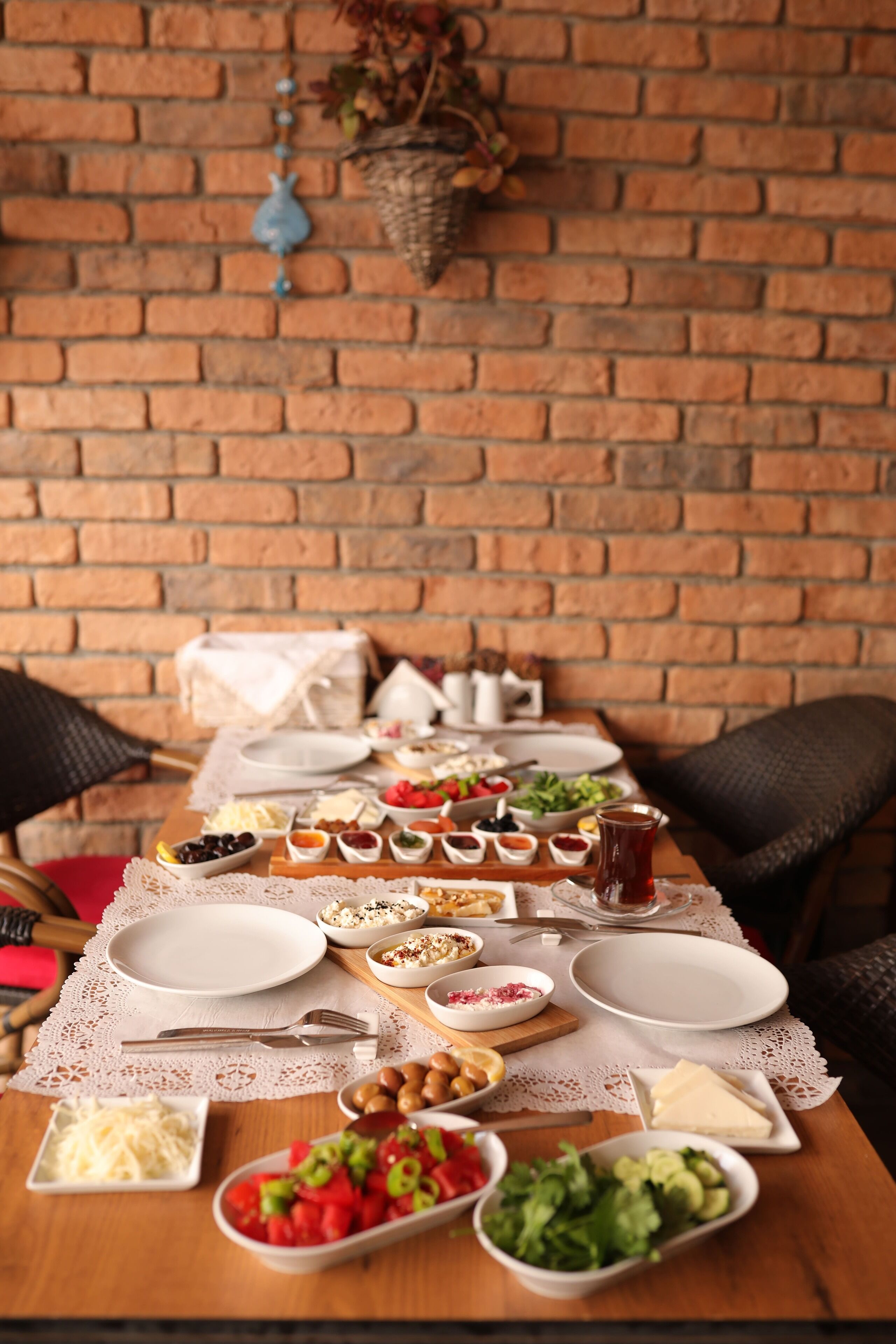 Uma mesa de jantar posta com diversos pratos e utensílios, parede de tijolos ao fundo, e cadeiras de vime.