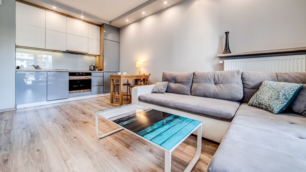 A modern living room with a grey sofa, a wooden coffee table, and a kitchen area with white cabinets and a dining table. - Rint - Centrum Kaczorowskiego (Bialystok)
