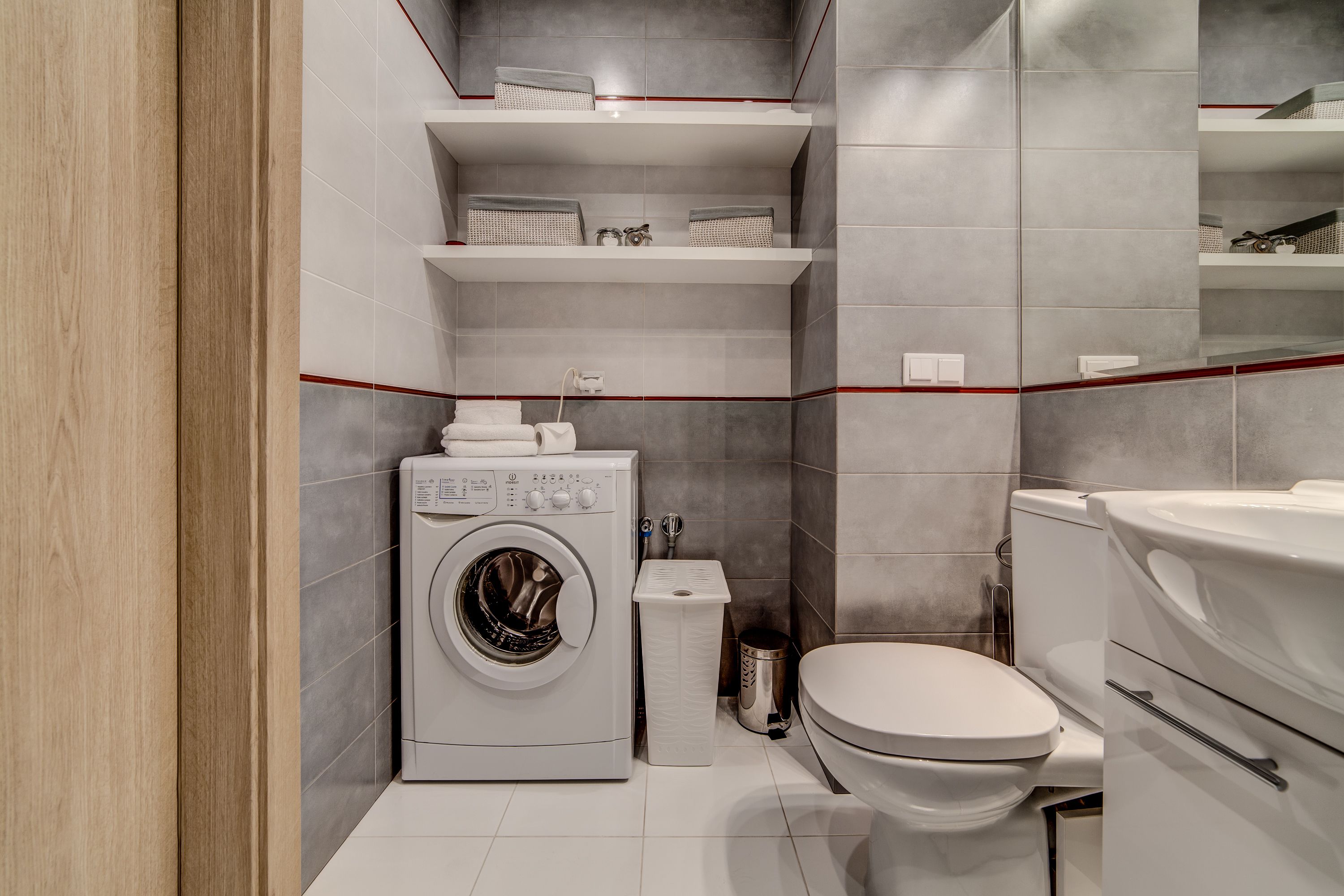 A compact bathroom with a washing machine, toilet, and shelves with toiletries.
