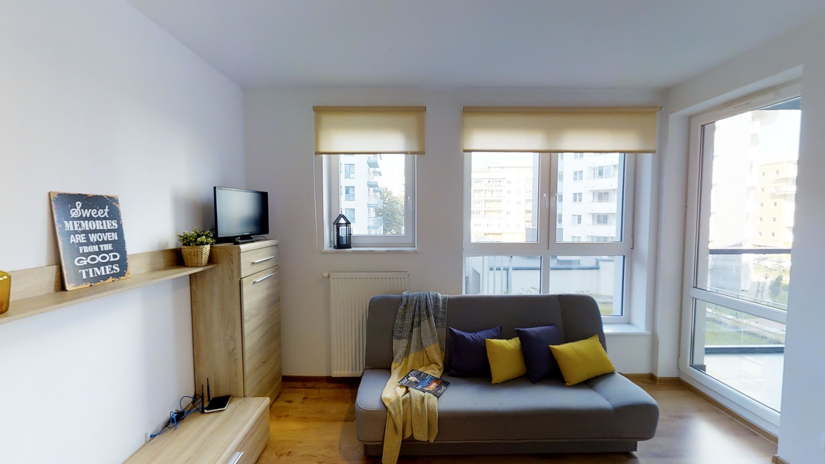 A modern living room with a grey sofa, a flat-screen TV on a wooden cabinet, a sign on the wall, and large windows with blinds.
