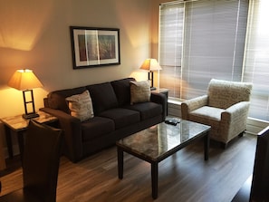 A living room with a dark sofa, two armchairs, a coffee table, and two lamps. - Weichert Suites at The Bartlett (Arlington)