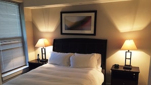 A neatly made bed with white linens, two bedside tables with lamps, a framed picture on the wall, and a window with blinds. - Weichert Suites at The Bartlett (Arlington)