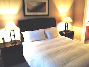 A neatly made bed with white linens, two bedside lamps, and a framed picture on the wall. - Weichert Suites at The Bartlett (Arlington)