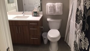 A bathroom with a white sink, wooden cabinets, a toilet, and a shower curtain with a floral pattern. - Weichert Suites at The Bartlett (Arlington)