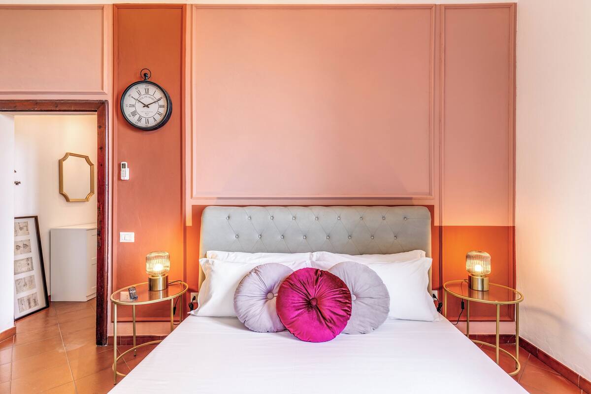 A neatly made bed with white linens and decorative pillows, a wall clock, and two bedside lamps on a bedside table.