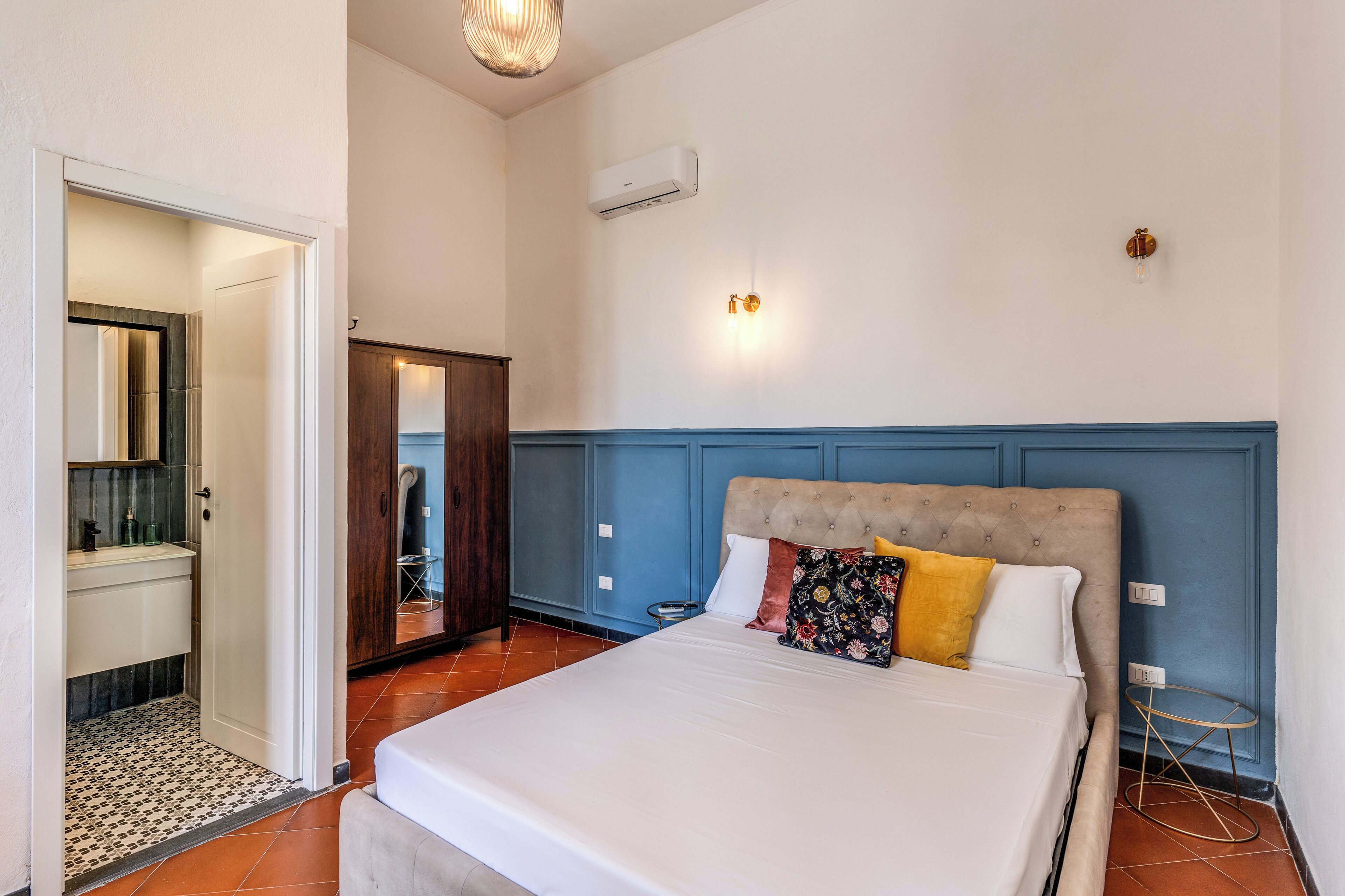 A bedroom with a large bed, a wooden wardrobe, a bathroom with a mirror, and a small round table.