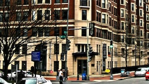 A multi-story building with a red brick facade and beige trim, featuring a corner entrance and multiple windows. - Weichert Suites at Thomas Circle (Washington)