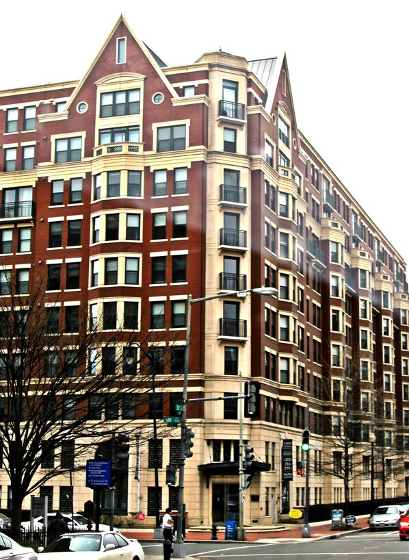 A multi-story building with a red brick facade and beige trim, featuring a corner entrance and multiple windows.