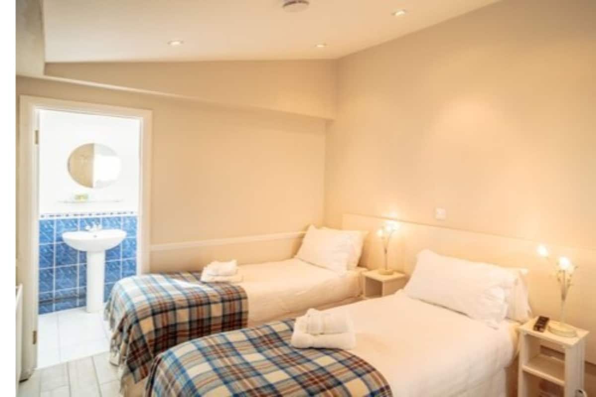 Une chambre d’hôtel avec deux lits, une salle de bain équipée d’un lavabo et d’un miroir, et une table de chevet avec une lampe.