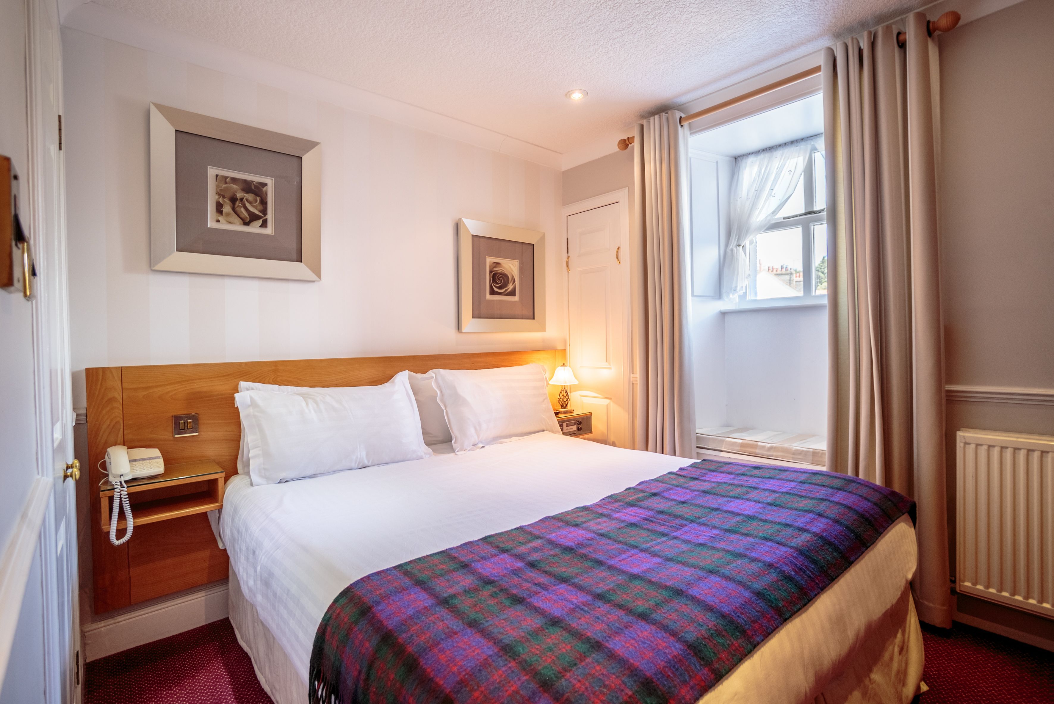 Habitación de hotel con una cama grande, una mesita de noche de madera con un teléfono, dos cuadros enmarcados en la pared, una ventana con cortinas y un radiador.