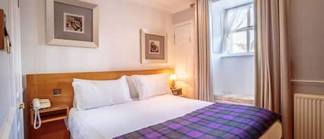 Habitación de hotel con una cama grande, una mesita de noche de madera con un teléfono, dos cuadros enmarcados en la pared, una ventana con cortinas y un radiador.