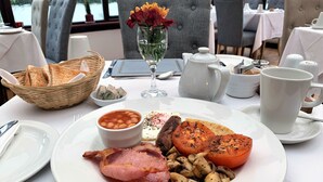Een ontbijttafel met een bord eieren, spek, champignons en tomaten, een glas water, een vaas met bloemen en een broodmand.