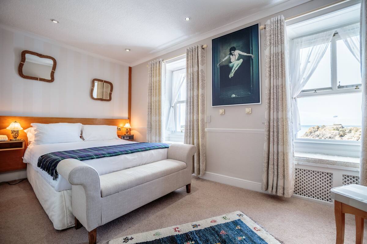 Une chambre à coucher avec un lit, des lampes de chevet, un banc, une fenêtre avec des rideaux et une affiche accrochée au mur.