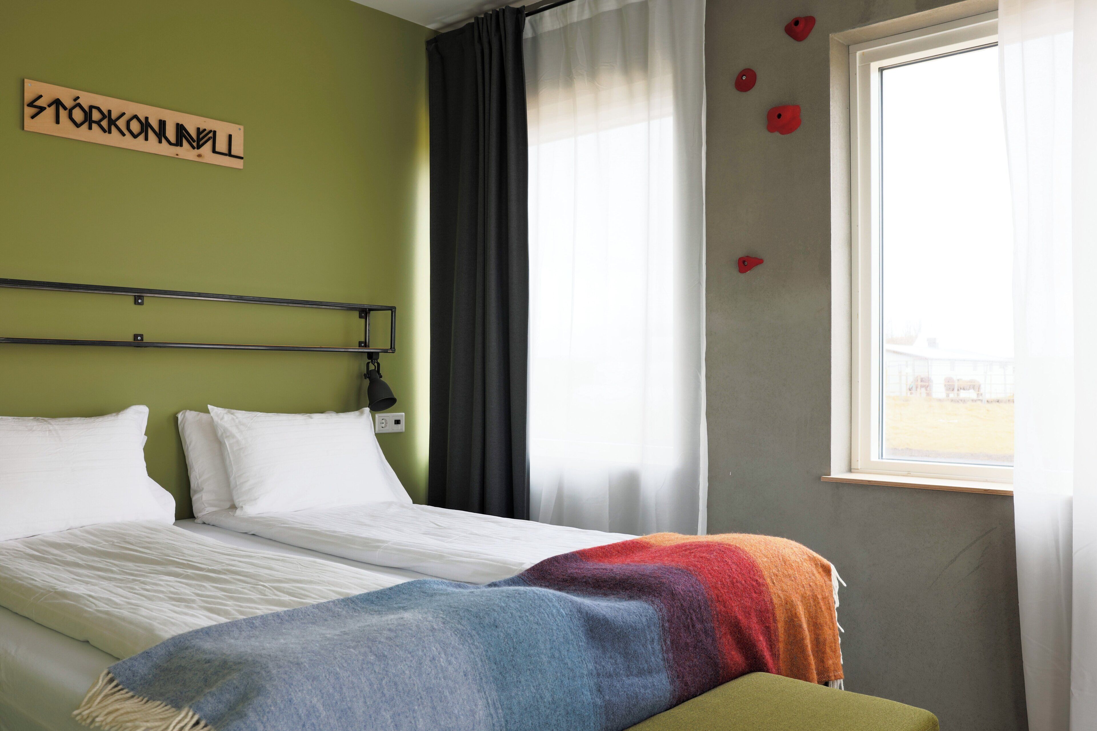 A hotel room with a bed, a bedside table, a lamp, a window with curtains, and a sign on the wall.