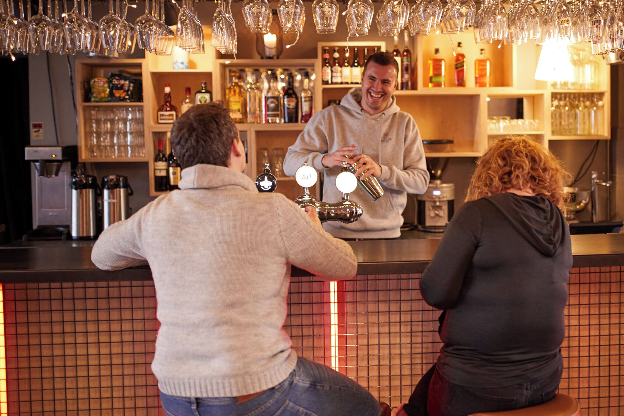 A bar area with a bartender preparing drinks, shelves with bottles, and a tiled wall.