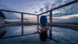 A person in a hot tub with a view of a sunset over a body of water and distant hills. - Midgard Base Camp (Rangárþing eystra)
