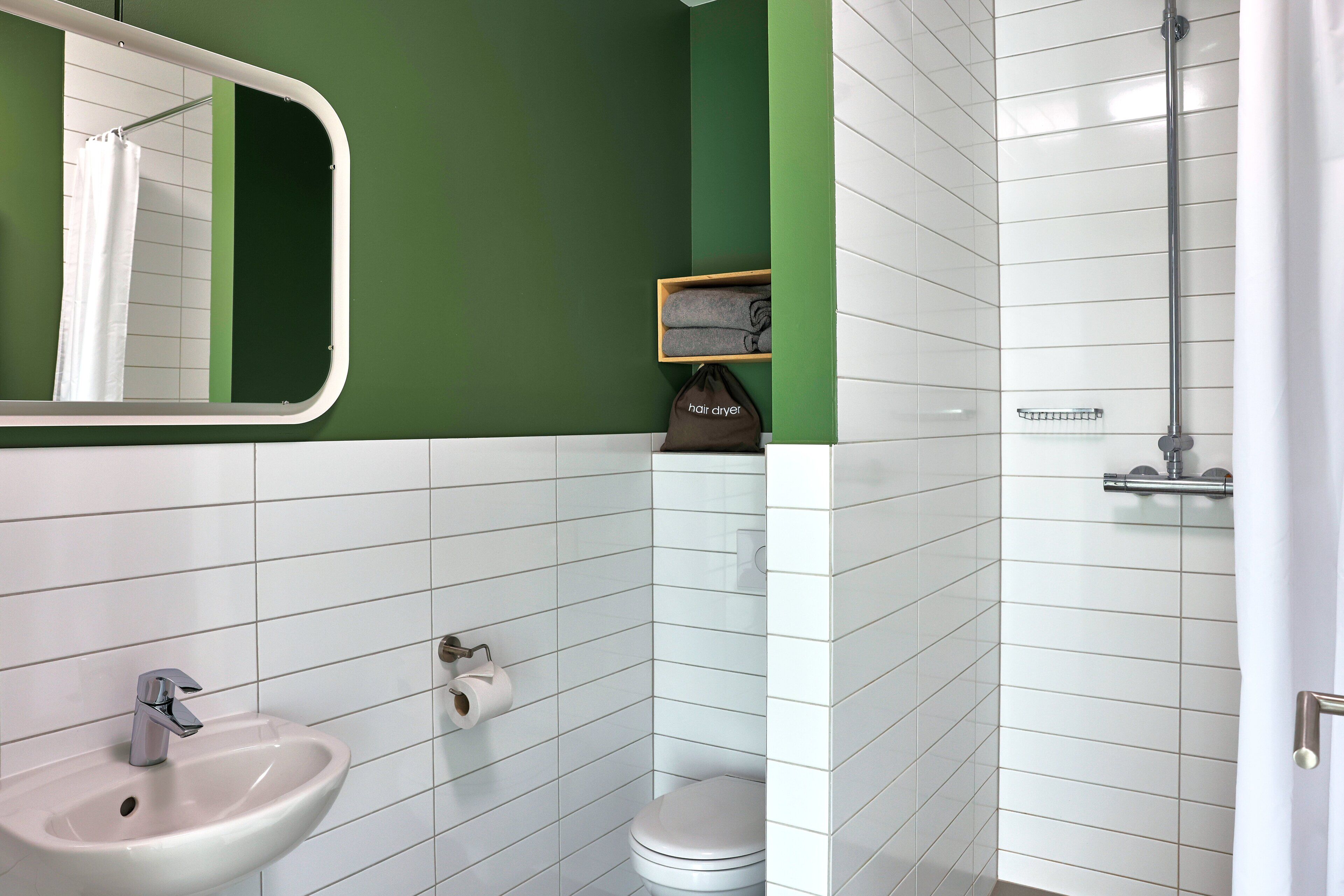 Baño con lavamanos, inodoro y ducha blancos. Hay un espejo sobre el lavamanos y un perchero con una toalla.