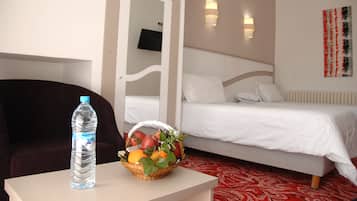 A hotel room with a bed, a bedside table with a water bottle and fruit basket, a chair, and a TV on the wall.