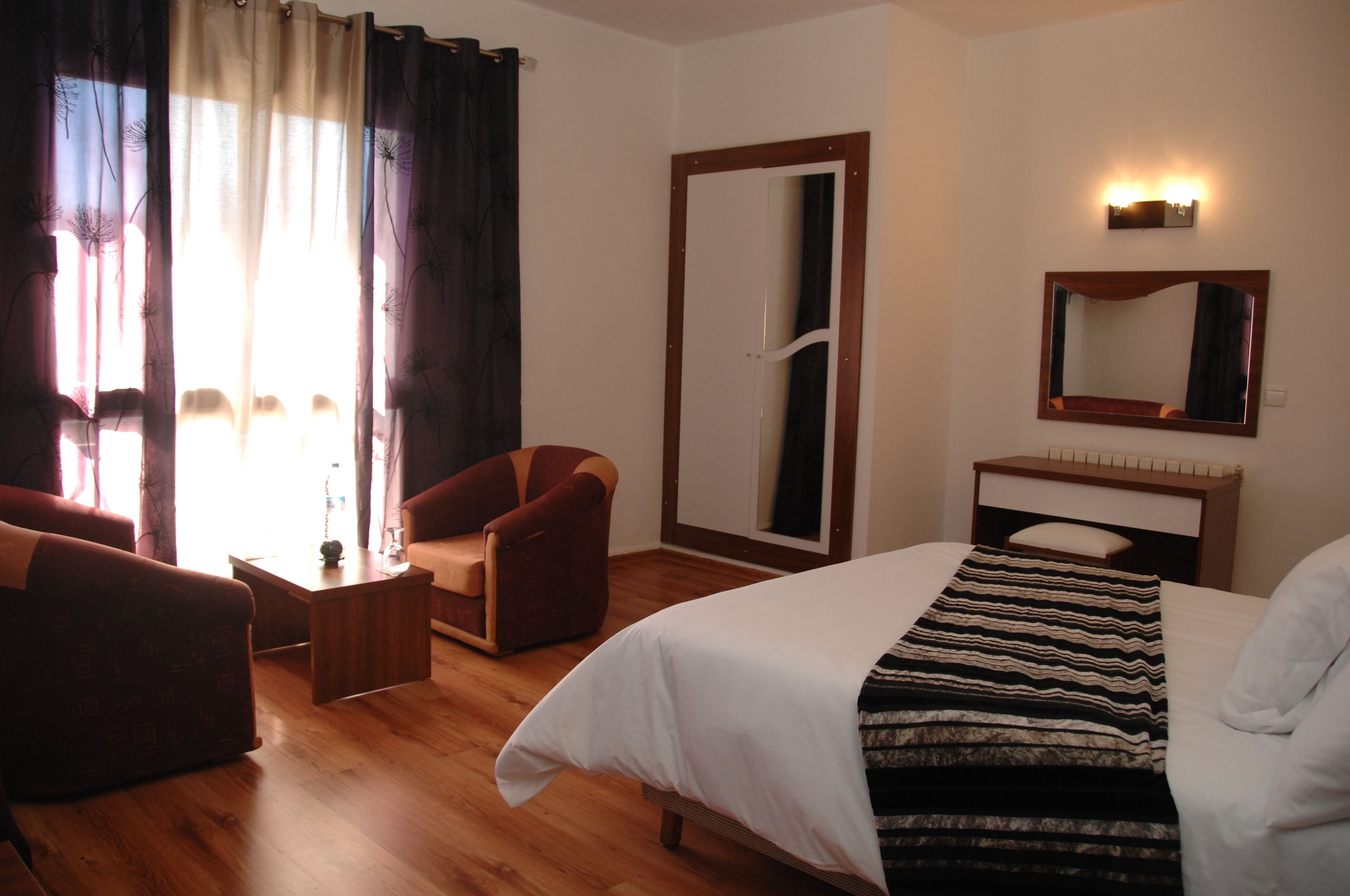 Ein Hotelzimmer mit Bett, Sessel, kleinem Tisch, Spiegel und einem Fenster mit Vorhängen.