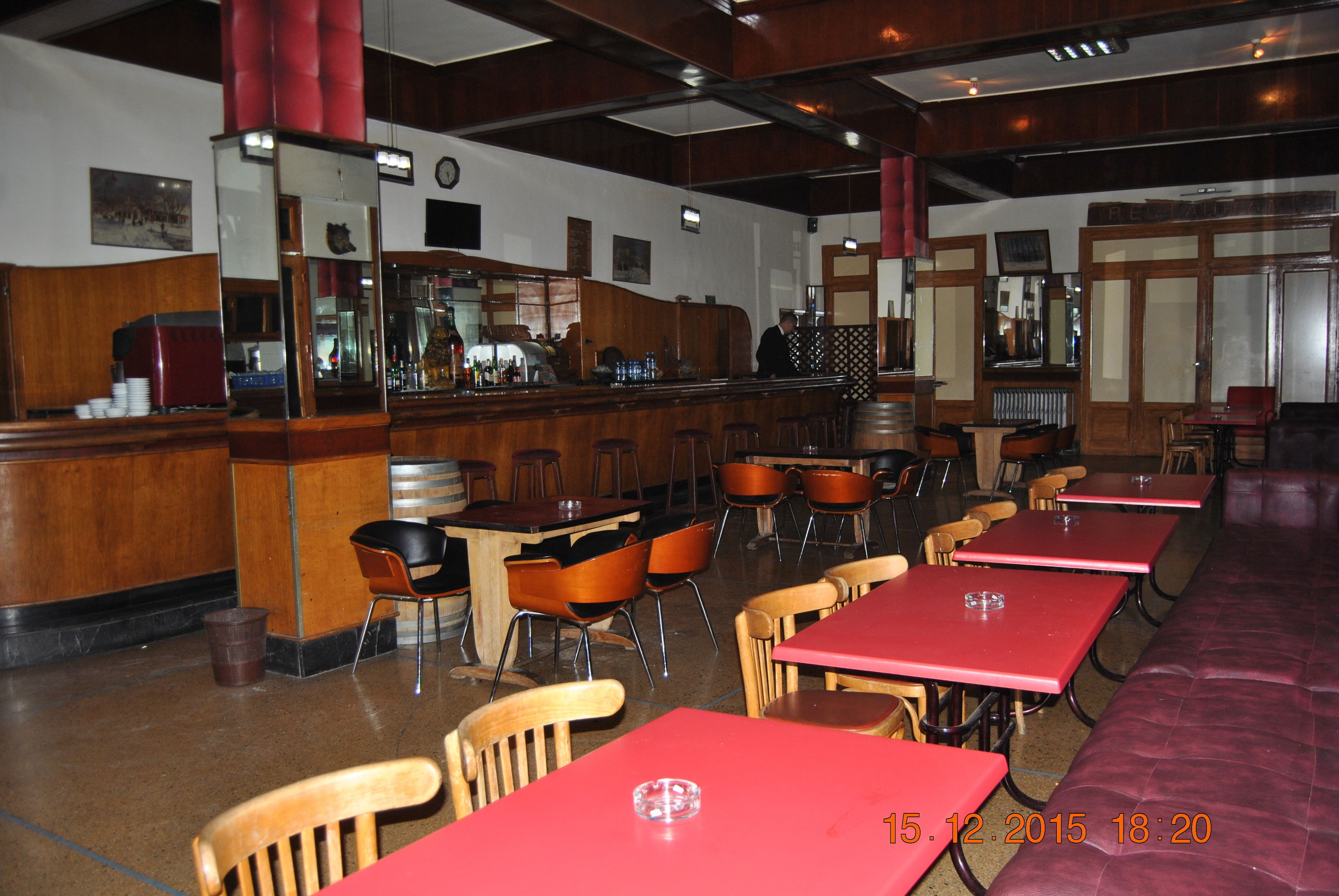 A restaurant interior with wooden tables and chairs, a bar area, and a large red table in the foreground.