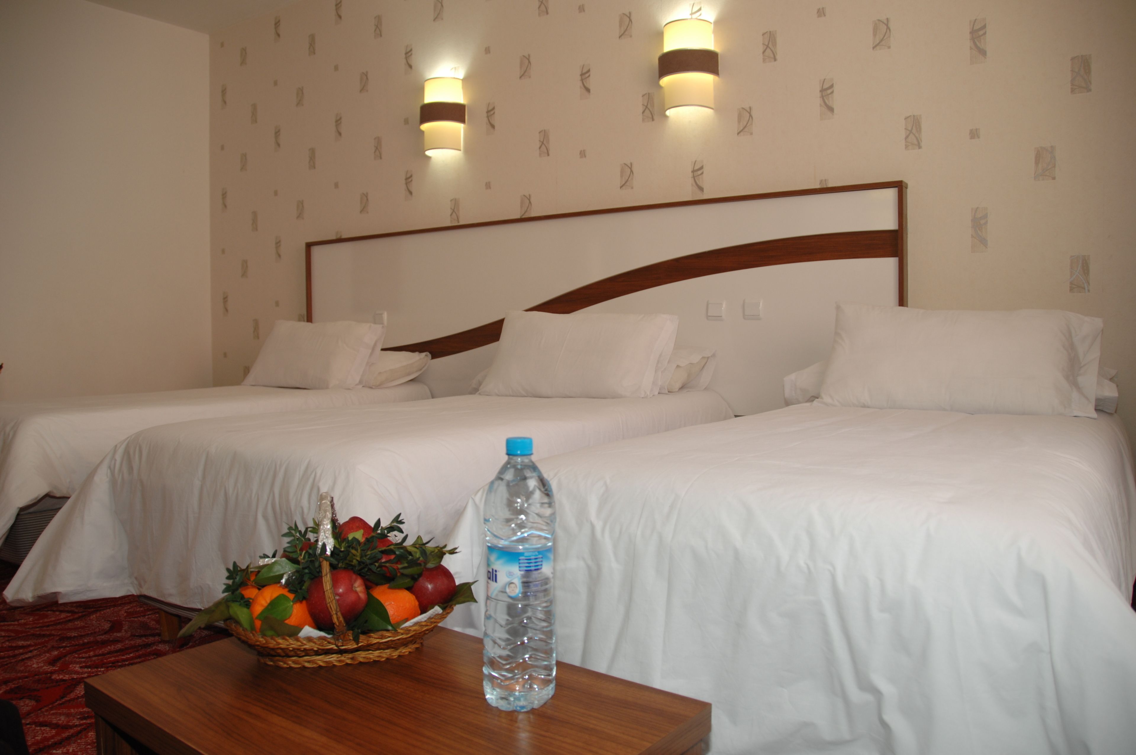 A hotel room with two beds, a wooden headboard, white bedding, a fruit basket, and a water bottle on a wooden table.