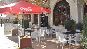 An outdoor seating area with white chairs and tables, a red Coca-Cola umbrella, and a stone building with arched windows.