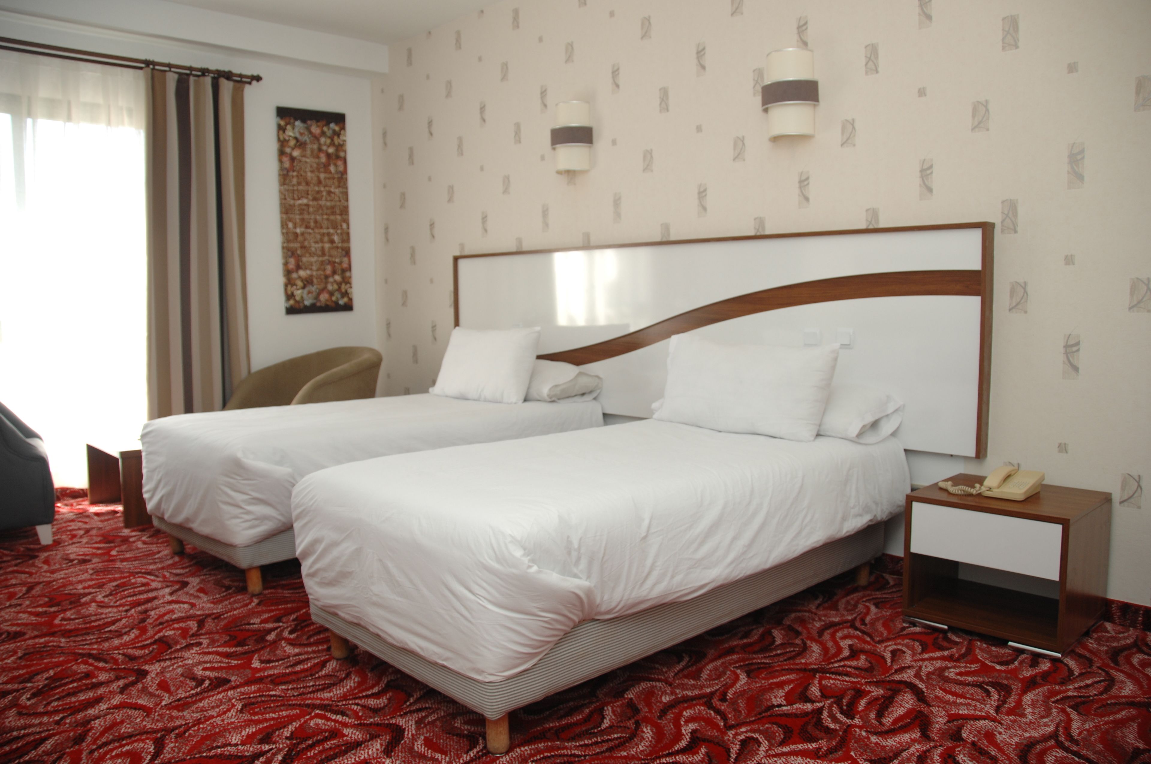 Une chambre d’hôtel avec deux lits, une moquette rouge à motifs, une chaise, une table de chevet avec un téléphone et un mur recouvert d’un papier peint à motifs.