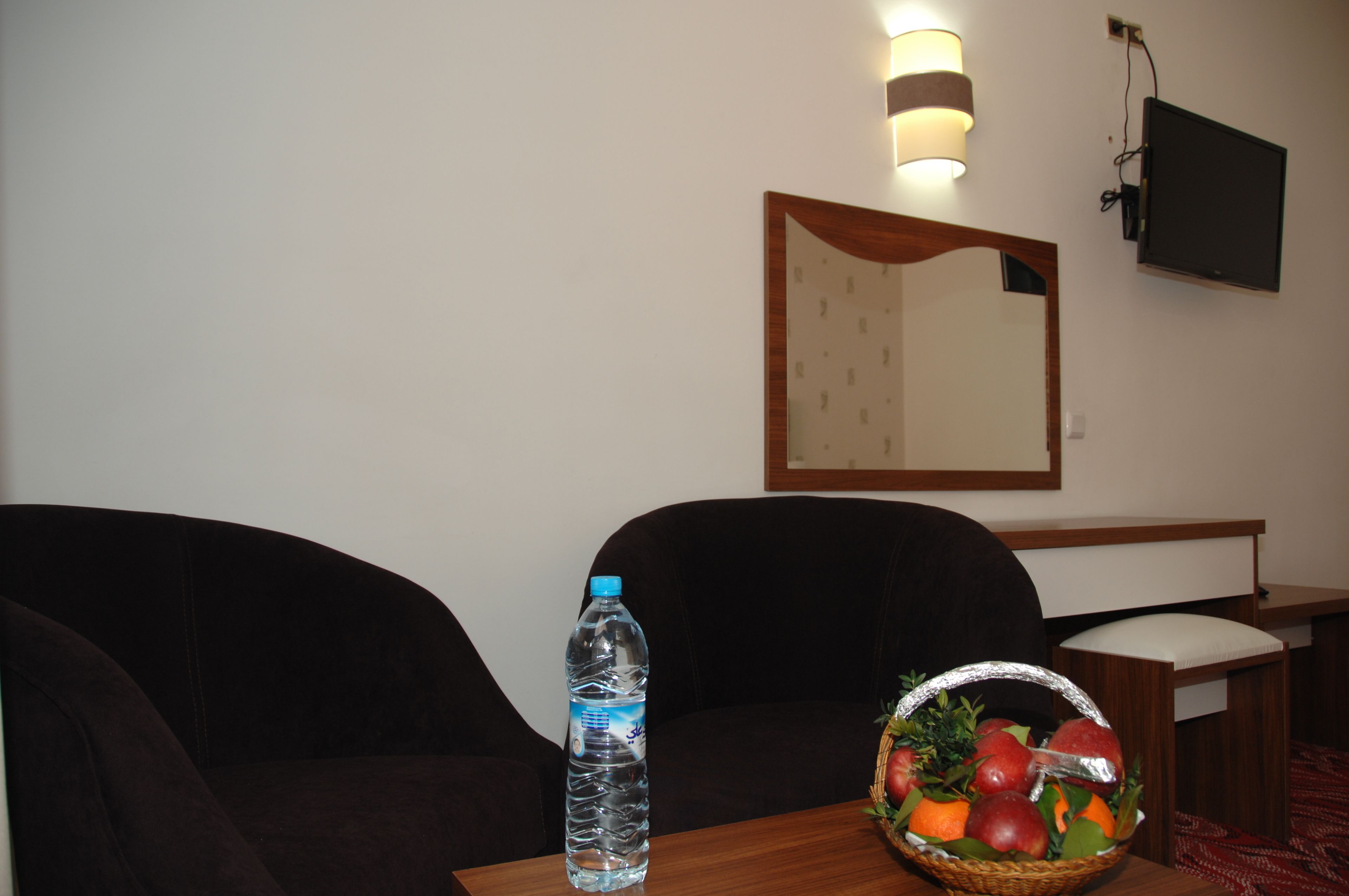 A hotel room with a dark brown sofa, a wooden table with a water bottle and fruit basket, a flat-screen TV mounted on the wall, and a mirror above the sofa.