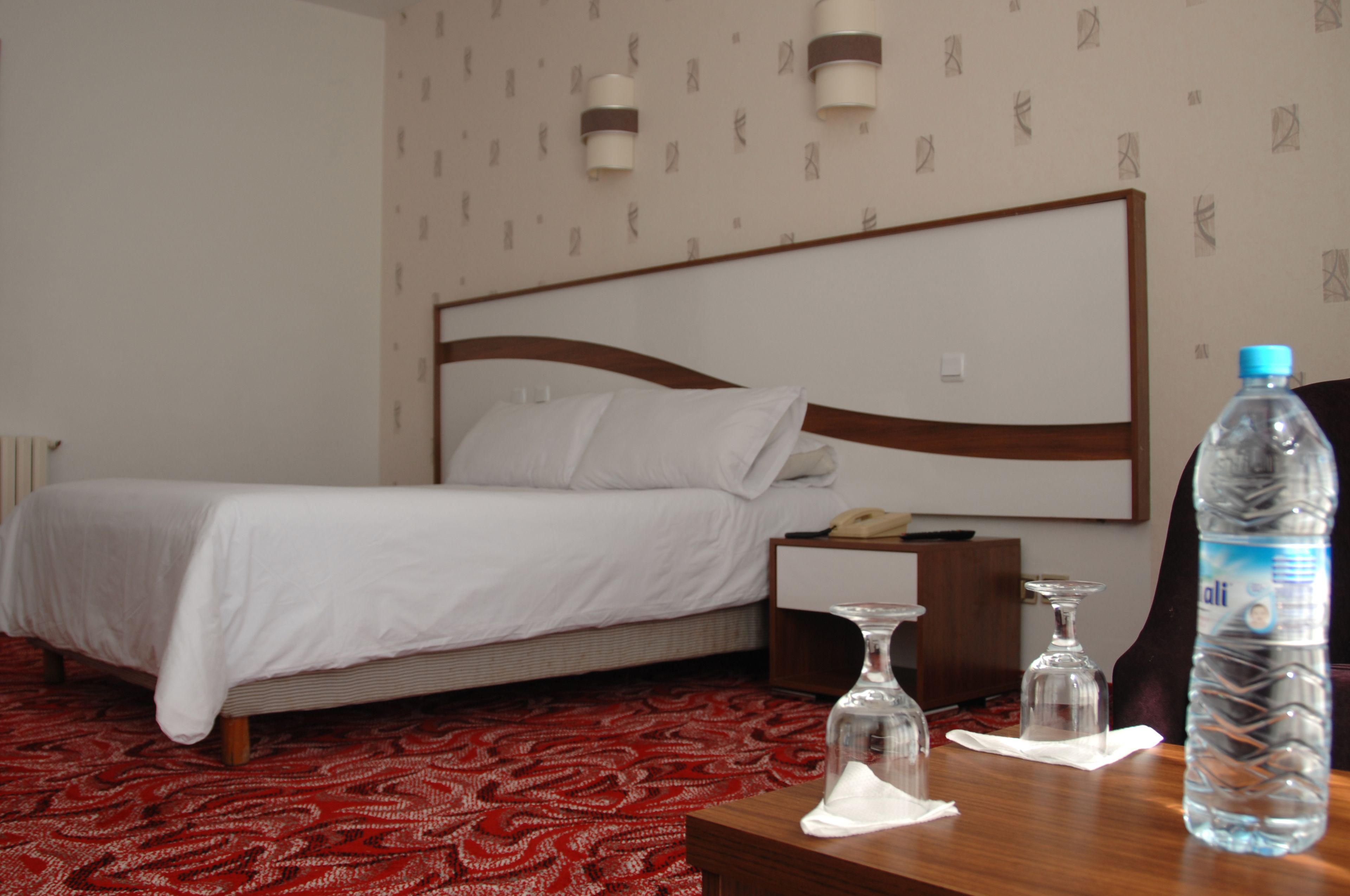 A hotel room with a bed, a wooden nightstand, a bedside table with a water bottle, a glass, and a telephone.