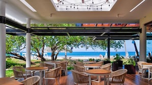 An outdoor seating area with tables and chairs, overlooking a beach with umbrellas and lounge chairs. - Azura Beach Resort - All Inclusive - Adults Only (Sámara)