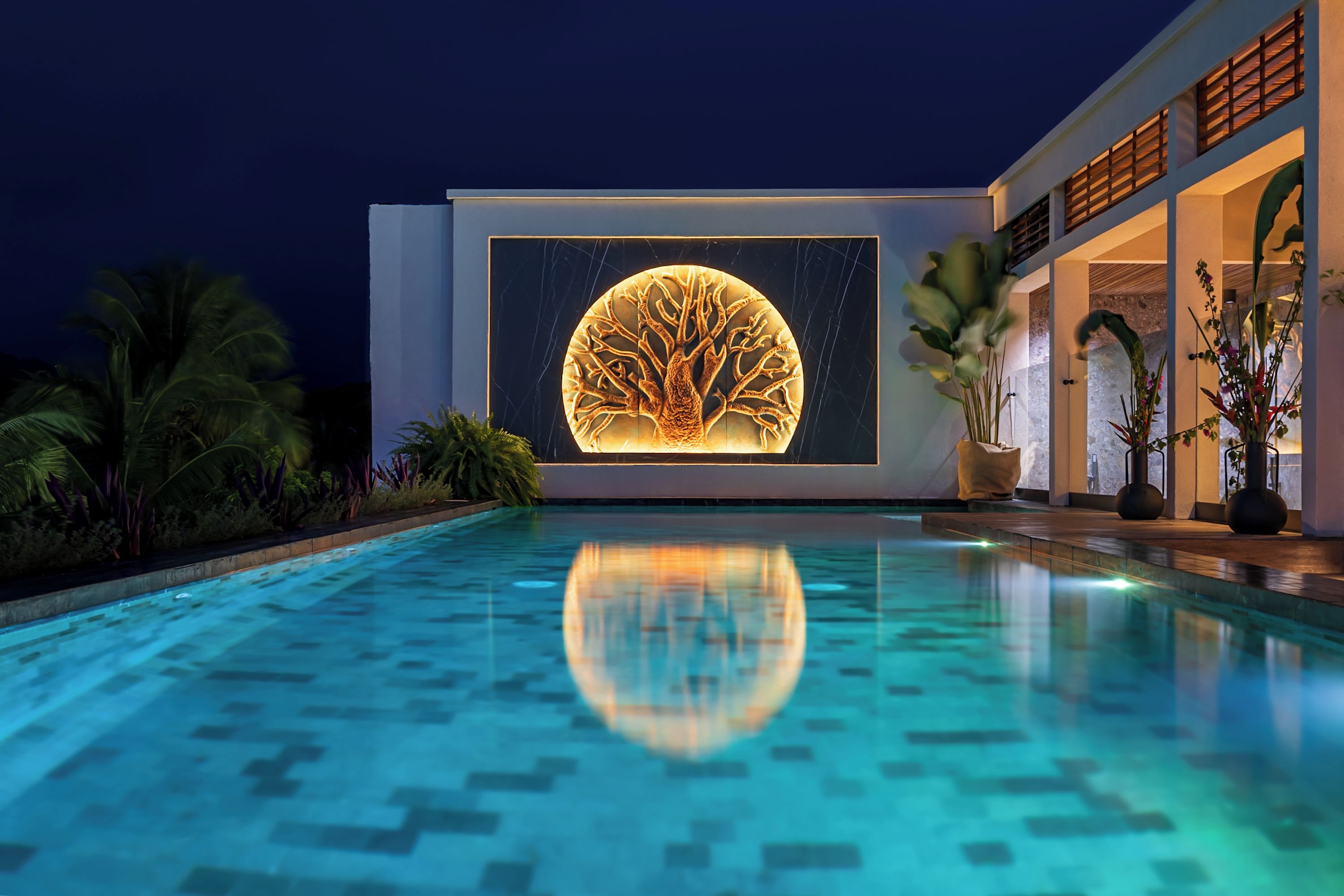 Un bâtiment moderne avec un bassin d’eau réfléchissant à l’avant, une grande sculpture d’arbre illuminée sur la façade et des plantes tropicales tout autour.