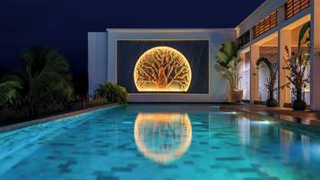 Un bâtiment moderne avec un bassin d’eau réfléchissant à l’avant, une grande sculpture d’arbre illuminée sur la façade et des plantes tropicales tout autour.